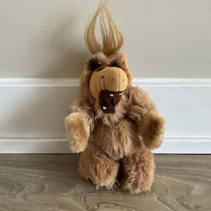 May include: A plush toy of the character Alf, featuring brown fur, a tan snout, and distinctive spiky hair. The toy has a brown patch on its mouth and is standing upright.
