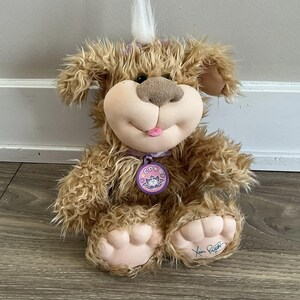 Puede incluir: Un juguete de peluche de perro marrón y blanco con la lengua rosa que sobresale. El perro tiene un collar morado con un corazón rosa y las letras "CPK" en él. El perro está sentado en un suelo de madera.