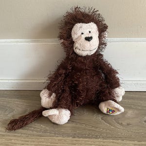 Puede incluir: Un mono de peluche marrón con cara, manos y pies blancos. El mono tiene una cola larga y está sentado en un suelo de madera. La cara del mono tiene una nariz negra en forma de corazón y una sonrisa cosida.