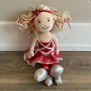 May include: A plush doll with blonde yarn hair featuring red highlights and a matching red bow. The doll wears a red velvet bodice with silver sequin trim, a layered red tulle skirt, a pearl necklace, and silver ballet slippers with ribbons.