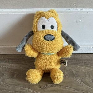 May include: A yellow plush toy dog with grey ears and a light blue collar. The toy has a white face with black eyes and a grey nose. The toy is sitting on a wooden floor. The toy has a tag that says "Goofy".