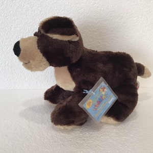 Puede incluir: Juguete de peluche de perro marrón y beige con una etiqueta de Webkinz adjunta. La etiqueta tiene un fondo azul con un borde blanco y presenta un perro de dibujos animados con un lazo rosa.