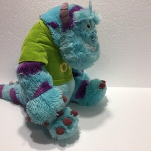 Op de afbeelding: Blauw en paars pluche monster speelgoed dat een groen shirt draagt met de letter "O" erop.