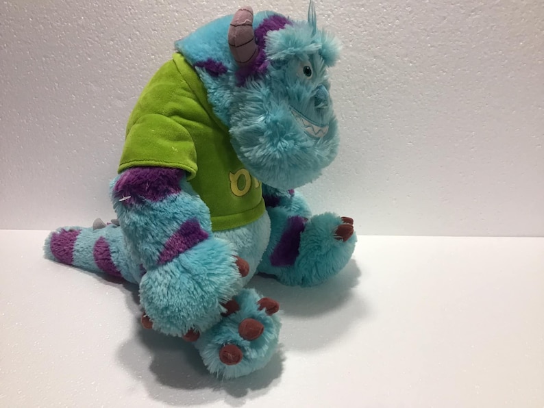 Op de afbeelding: Blauw en paars pluche monster speelgoed dat een groen shirt draagt met de letter "O" erop.