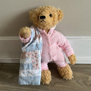 Puede incluir: Un oso de peluche con un pijama rosa con ribete blanco y sosteniendo una colcha de retazos azul y rosa.