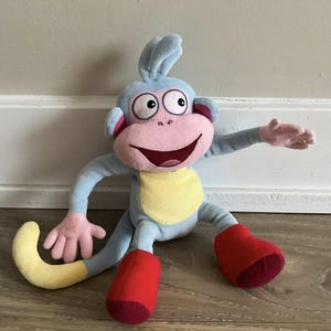 Puede incluir: Un mono de peluche azul y amarillo con la cara rosa y zapatos rojos. El mono tiene una gran sonrisa y está sentado en un suelo de madera.