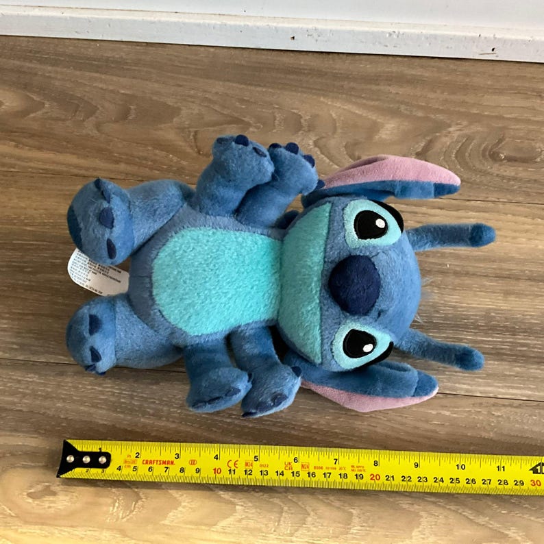 May include: A blue plush toy character with large ears, a light blue belly, and dark blue nose, positioned above a yellow measuring tape showing a length of approximately 11 inches from the top of the head to the bottom of the feet.