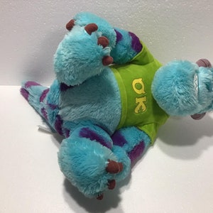 Op de afbeelding: Blauw en paars pluche speelgoed van een monster met een groen shirt met de tekst "OK" erop.