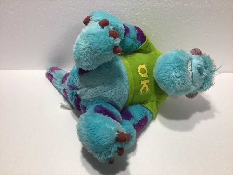 Op de afbeelding: Blauw en paars pluche speelgoed van een monster met een groen shirt met de tekst "OK" erop.