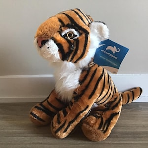 May include: A plush sitting tiger toy with orange and black stripes. The toy has a white chest and a tag that reads "animal planet plush sitting tiger 3+".
