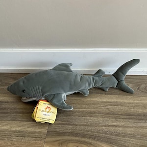 May include: A gray plush shark puppet with a detailed design, including fins, a tail, and a mouth. The puppet has a tag attached with the brand name "FOLKMANIS Puppets". The shark is lying on a wooden surface.