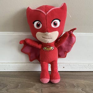 以下が含まれることがあります： ピンクのマスクと翼を持つスーパーヒーローのキャラクターの赤いぬいぐるみ。キャラクターは胸に金のエンブレムが付いた赤いスーツを着ています。