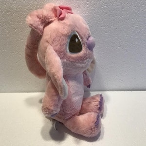 May include: Pink plush toy with a purple bow and brown eyes. The toy is sitting on a white surface.