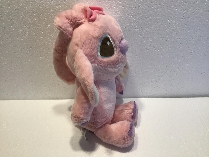 May include: Pink plush toy with a purple bow and brown eyes. The toy is sitting on a white surface.