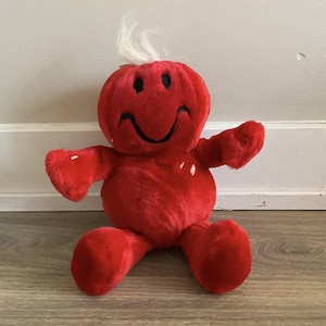 May include: A plush red character with a smiling face, black eyes, and a white tuft of hair. The stuffed toy has arms and legs, and is sitting on a wooden floor. The toy is a cheerful, friendly design.