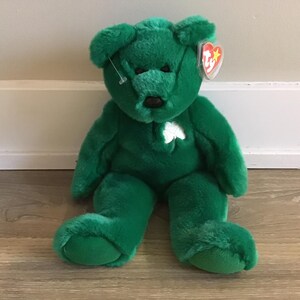May include: Green plush teddy bear with a white shamrock on its chest. The bear has a red and white Ty tag attached to its ear.