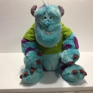 Op de afbeelding: Een blauw en paars pluche speelgoed van het personage Sulley uit de film Monsters Inc. Het speelgoed draagt een groen shirt met een gele zon.