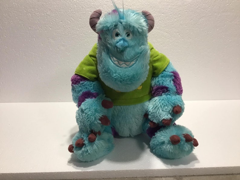 Op de afbeelding: Een blauw en paars pluche speelgoed van het personage Sulley uit de film Monsters Inc. Het speelgoed draagt een groen shirt met een gele zon.
