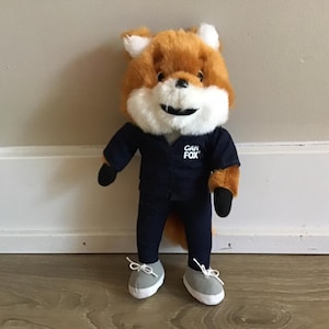 Pu&ograve; includere: Un peluche a forma di volpe con pelliccia arancione e bianca, che indossa un'uniforme blu navy con la scritta "CAR FOX" sul petto. La volpe ha scarpe da ginnastica grigie con lacci bianchi e guanti neri.