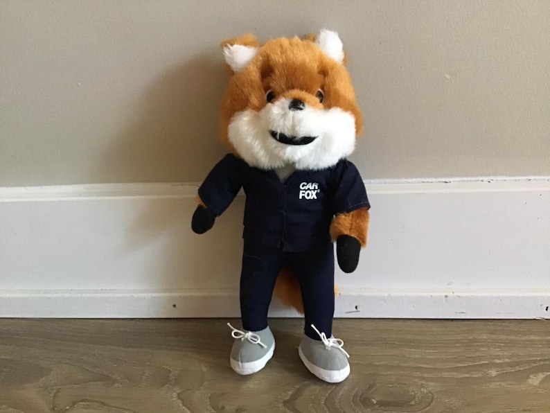 Pu&ograve; includere: Un peluche a forma di volpe con pelliccia arancione e bianca, che indossa un'uniforme blu navy con la scritta "CAR FOX" sul petto. La volpe ha scarpe da ginnastica grigie con lacci bianchi e guanti neri.