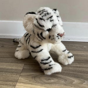 Op de afbeelding: Een wit pluche tijgerwelpje met zwarte strepen. De tijger heeft een roze neus en zit op een houten vloer.