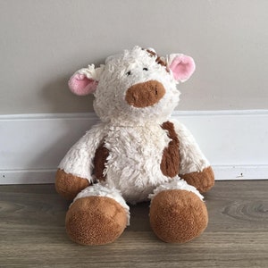 Pu&ograve; includere: Un giocattolo di peluche di mucca bianco e marrone con orecchie rosa. La mucca &egrave; seduta su un pavimento di legno.