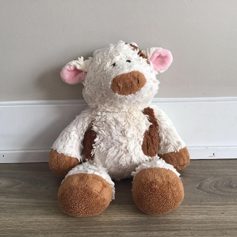 Pu&ograve; includere: Un giocattolo di peluche di mucca bianco e marrone con orecchie rosa. La mucca &egrave; seduta su un pavimento di legno.