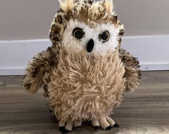 May include: A plush owl toy with a fluffy, textured body in shades of brown, tan, and white. The owl has large, round eyes, a black beak, and small, pointed ear tufts. The toy is standing on a wooden floor.