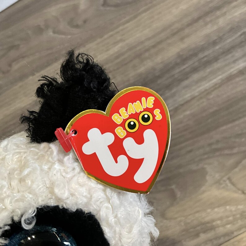 May include: A white and black fuzzy Beanie Boo with a red and gold heart-shaped tag that says "Beanie Boos ty".