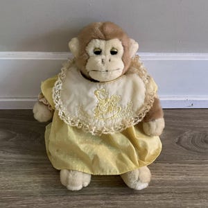 May include: A soft toy monkey with a light brown face and fur, dressed in a yellow dress with white polka dots and a cream-coloured bib with lace trim. The bib features a yellow embroidered design.