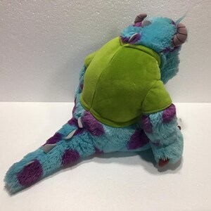 Op de afbeelding: Een blauw en paars pluche speelgoed van een monster met een groen shirt. Het monster heeft drie ogen en een grote mond.