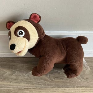 Pode incluir: Um urso de pelúcia com corpo marrom, rosto creme e marcas marrons escuras ao redor dos olhos. O urso tem orelhas vermelhas, nariz preto e boca sorrindo. O brinquedo foi projetado para se assemelhar a um urso de desenho animado.
