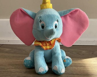 May include: A blue plush toy elephant with pink ears, a yellow hat, and a colorful collar. The elephant is sitting with its legs crossed and has a friendly expression.