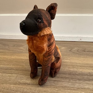 May include: A plush dog toy featuring a brindle patterned coat with dark brown and black stripes, a solid black muzzle, and a soft, textured chest area in a warm tan color. The stuffed animal is in a seated position with upright ears.