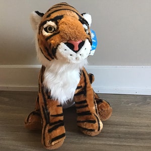 May include: A plush tiger toy with orange and black stripes, white fur on its chest, and a pink nose. The toy is sitting on a wooden floor.