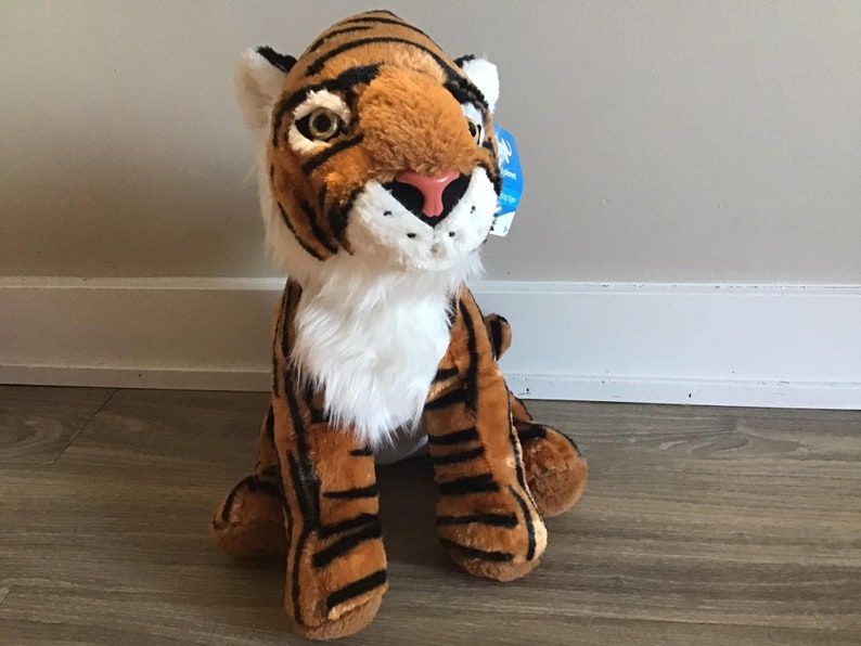 May include: A plush tiger toy with orange and black stripes, white fur on its chest, and a pink nose. The toy is sitting on a wooden floor.