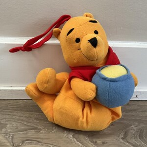 May include: A plush Winnie the Pooh toy in a seated position, holding a blue pot of honey. The bear is yellow with a red shirt and black eyes. A red ribbon is attached to the top of the bear's head.