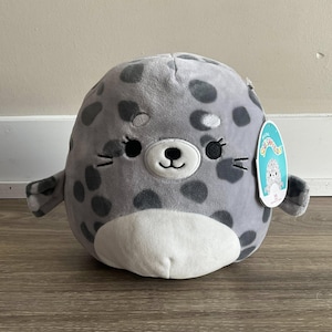 Puede incluir: Una foca de peluche gris y blanca con ojos negros y una nariz blanca. La foca tiene un patrón de manchas y sostiene una etiqueta que dice "Squishmallow".