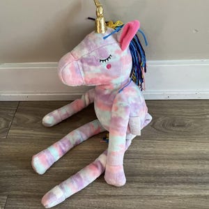 May include: A plush unicorn toy in pastel tie-dye colors. The unicorn has a gold horn, pink ears, and blue and yellow mane. The unicorn is sitting with its legs extended. The unicorn has closed eyes and a small red dot on its cheek.