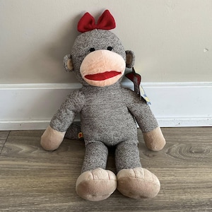 May include: A gray sock monkey plush toy with a red bow on its head. The monkey has a tan face, hands, and feet, and a red mouth. The toy is standing upright against a white wall and wooden floor.