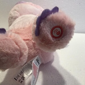 May include: Pink and purple plush toy with a Disney logo and a small button on the foot. The toy is lying on its back with its feet in the air.