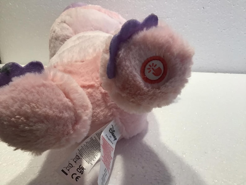 May include: Pink and purple plush toy with a Disney logo and a small button on the foot. The toy is lying on its back with its feet in the air.