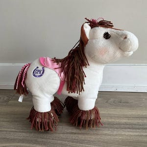 May include: White plush horse with brown yarn mane and tail. The horse has a pink saddle with a purple horseshoe logo that says "GPK".