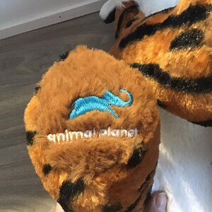 May include: Close-up of a brown and black plush tiger with a blue embroidered animal planet logo on the paw.