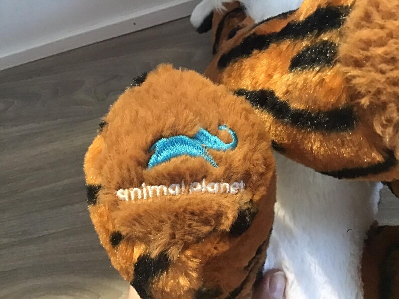 May include: Close-up of a brown and black plush tiger with a blue embroidered animal planet logo on the paw.