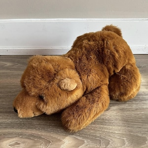 May include: A brown plush bear toy with a soft, furry texture. The bear is lying down, with its head and paws visible. The toy has small, rounded ears and a small tail. The bear is a warm, inviting colour.