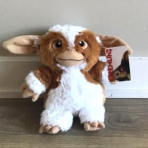 May include: A plush Gremlins toy with brown and white fur, large ears, and a tag that reads "Gremlins". The toy is standing on a wooden floor.