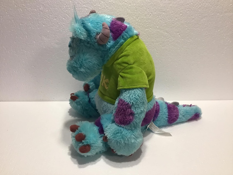 Op de afbeelding: Een blauw en paars pluche speelgoed van het personage Sulley uit de film Monsters Inc. Het speelgoed draagt een groen shirt met het woord "Mike" erop.