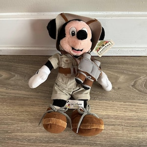 May include: A plush Mickey Mouse toy dressed in a tan safari outfit with a matching hat, brown boots with gray laces, and a pair of gray and brown binoculars hanging around the neck. A tag attached to the ear reads Animal Kingdom, and a tag reads Walt Disney World.