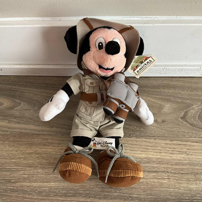 May include: A plush Mickey Mouse toy dressed in a tan safari outfit with a matching hat, brown boots with gray laces, and a pair of gray and brown binoculars hanging around the neck. A tag attached to the ear reads Animal Kingdom, and a tag reads Walt Disney World.